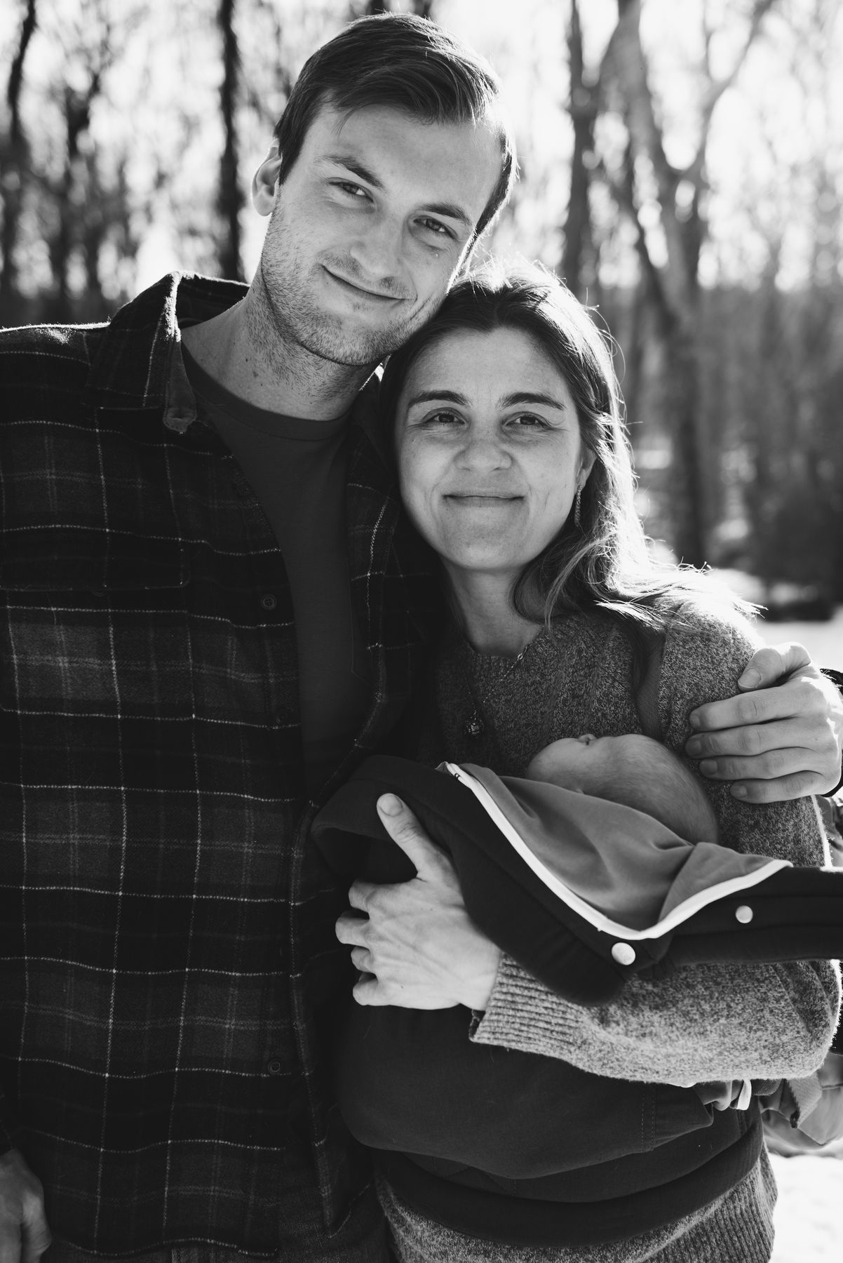 New parents smiling, baby carrier on his chest, in black and white